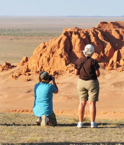 Two people photograph a rocky, arid landscape with reddish-orange formations under a clear sky.