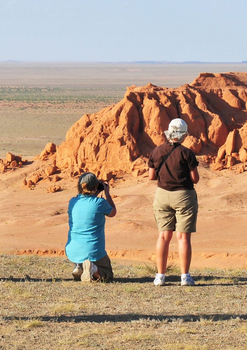 Two people photograph a rocky, arid landscape with reddish-orange formations under a clear sky.