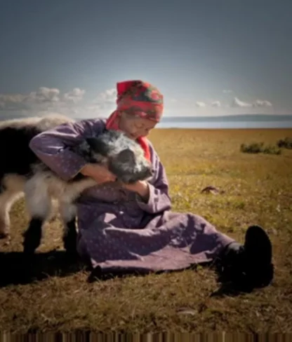 Person in a purple outfit sitting on grass, holding a black and white animal. They are wearing a red patterned headscarf. The background shows a plain with a distant body of water.