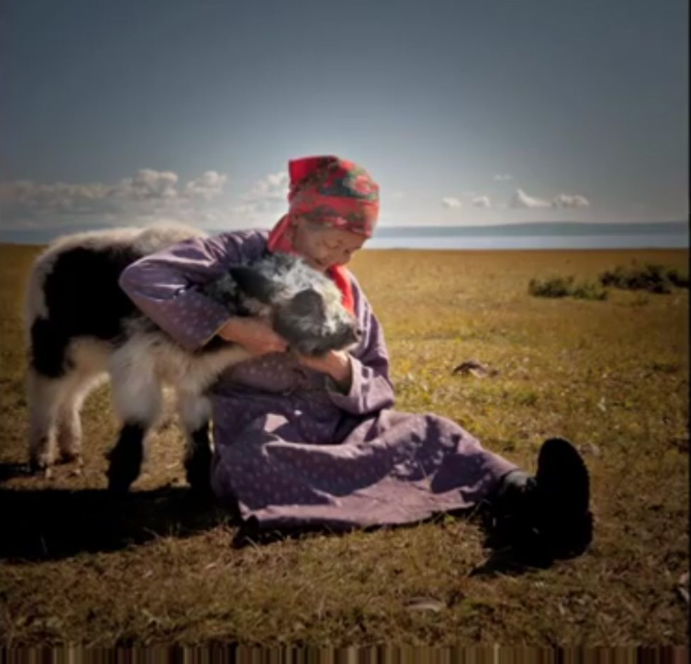 Person in a purple outfit sitting on grass, holding a black and white animal. They are wearing a red patterned headscarf. The background shows a plain with a distant body of water.