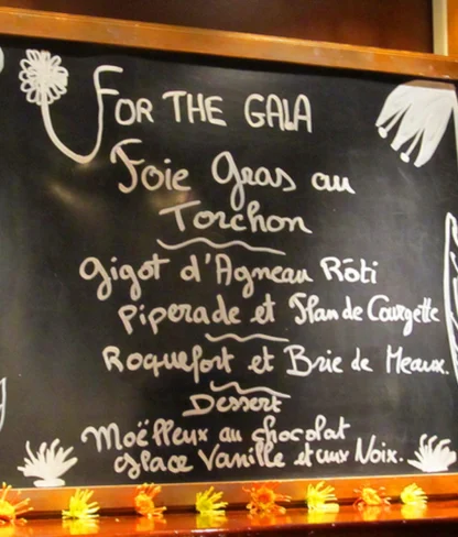 Chalkboard menu for a gala featuring foie gras, roasted lamb, piperade, Roquefort and Brie, and a chocolate dessert. Bottles of wine are placed nearby.