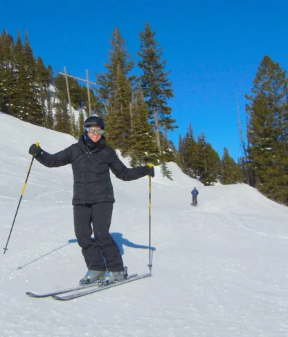 Person skiing down a snowy slope with evergreen trees in the background under a clear blue sky.