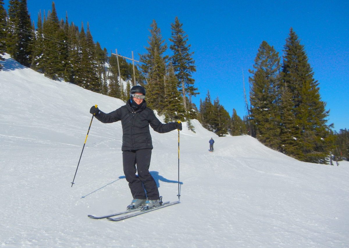 Person skiing down a snowy slope with evergreen trees in the background under a clear blue sky.