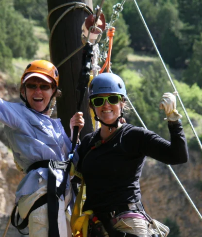 Two people wearing helmets and harnesses are smiling and posing on a zip line against a backdrop of trees and rocky cliffs.