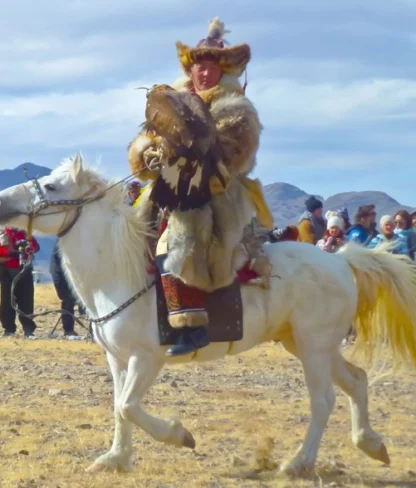 A person in traditional attire rides a white horse while holding a large bird, with a crowd and mountainous landscape in the background.