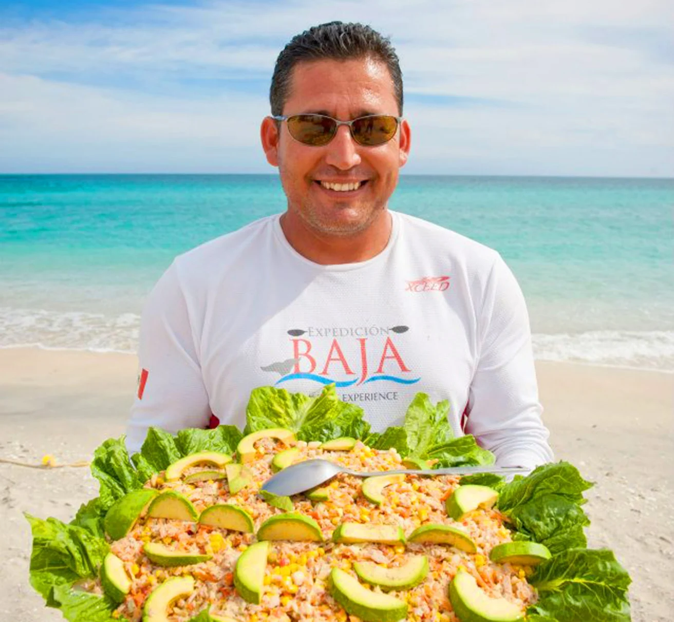 A man in sunglasses stands on a beach, holding a large platter of food topped with avocado slices and lettuce.