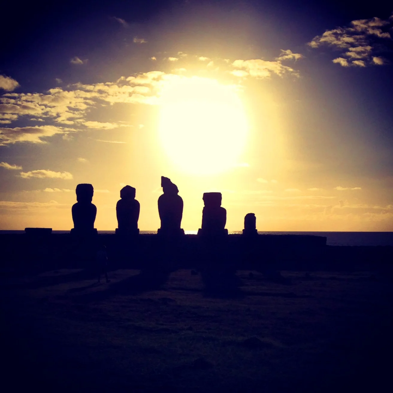 Silhouettes of Easter Island moai statues against a setting sun and a partly cloudy sky.