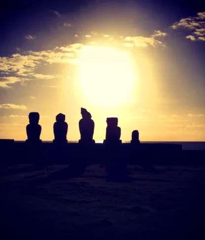 Silhouettes of Easter Island moai statues against a setting sun and a partly cloudy sky.