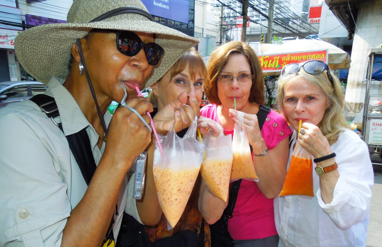 Four women drink beverages from plastic bags with straws on a busy street, with shops and signs in the background.