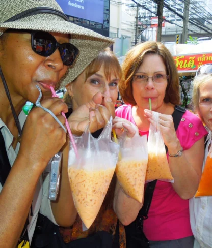 Four women drink beverages from plastic bags with straws on a busy street, with shops and signs in the background.