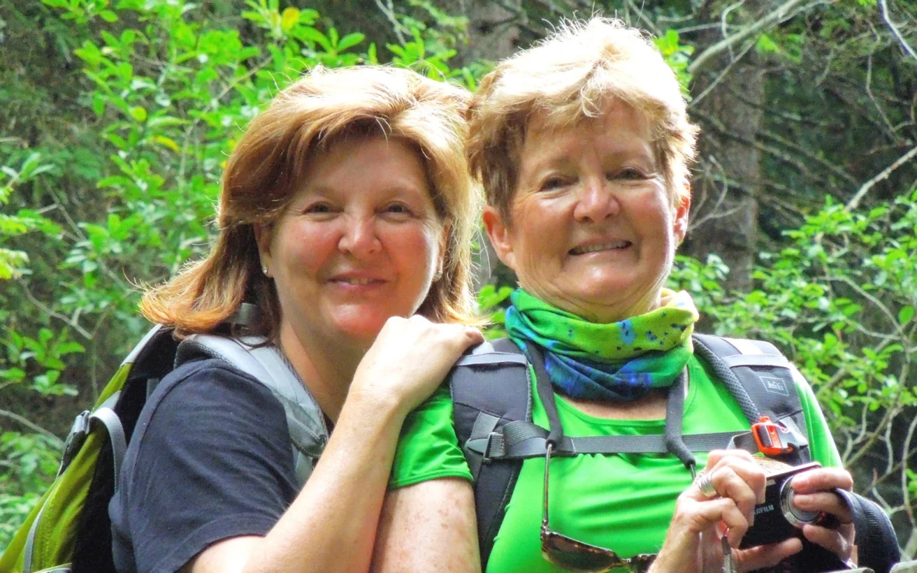 Two people wearing backpacks and bright clothing smiling outdoors in a green forested area. One holds a camera.