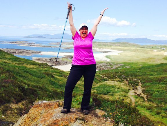 Person standing on a rock with arms raised, holding a walking stick, overlooking a green landscape and coastline under a clear blue sky.