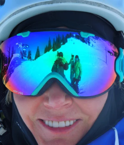 A person wearing mirrored ski goggles captures a snowy mountain reflection with another person waving. They are dressed in winter gear, including a helmet.