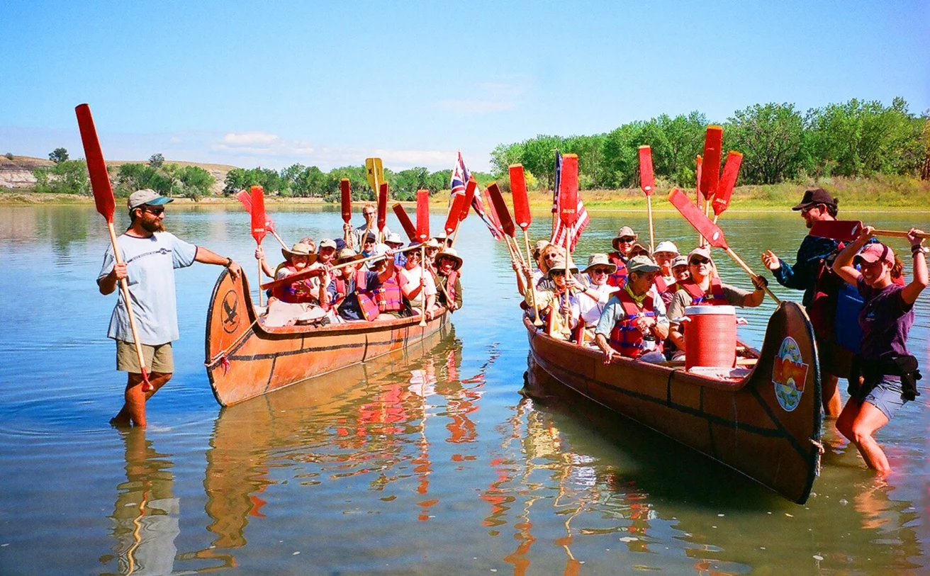 Two groups of people in canoes raise paddles on a calm river, guided by two instructors. The scene is bright and sunny with trees in the background.