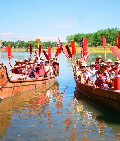 Two groups of people in canoes raise paddles on a calm river, guided by two instructors. The scene is bright and sunny with trees in the background.