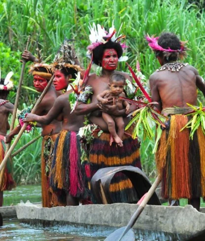 A group of people in traditional attire paddles a wooden canoe on a river surrounded by dense greenery.