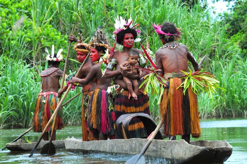 A group of people in traditional attire paddles a wooden canoe on a river surrounded by dense greenery.