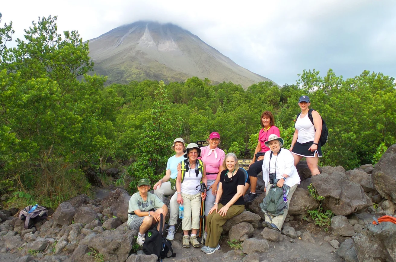 A group of hikers, wearing casual outdoor attire, pose on rocky terrain with a lush green backdrop. A large mountain is visible in the background under a cloudy sky.