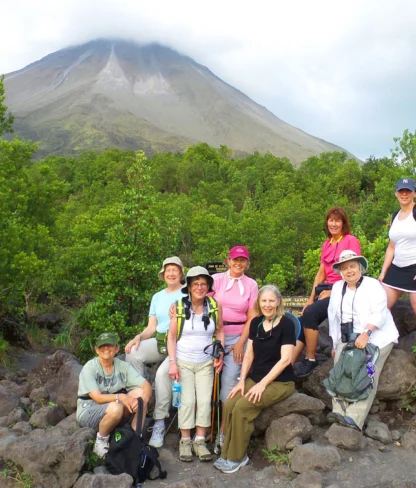 A group of hikers, wearing casual outdoor attire, pose on rocky terrain with a lush green backdrop. A large mountain is visible in the background under a cloudy sky.