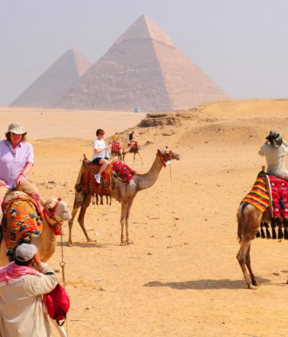 Tourists ride camels near the Pyramids of Giza, guided by locals in traditional clothing, with a clear sky overhead.