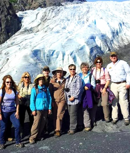 A group of people stand in front of a large glacier, posing for a photo. They are dressed in hiking gear and are on rocky terrain under sunny skies.