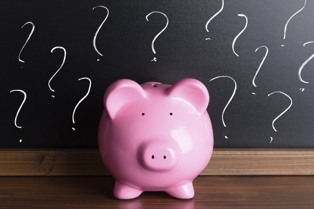 A pink piggy bank sits on a wooden surface in front of a blackboard with drawn question marks.