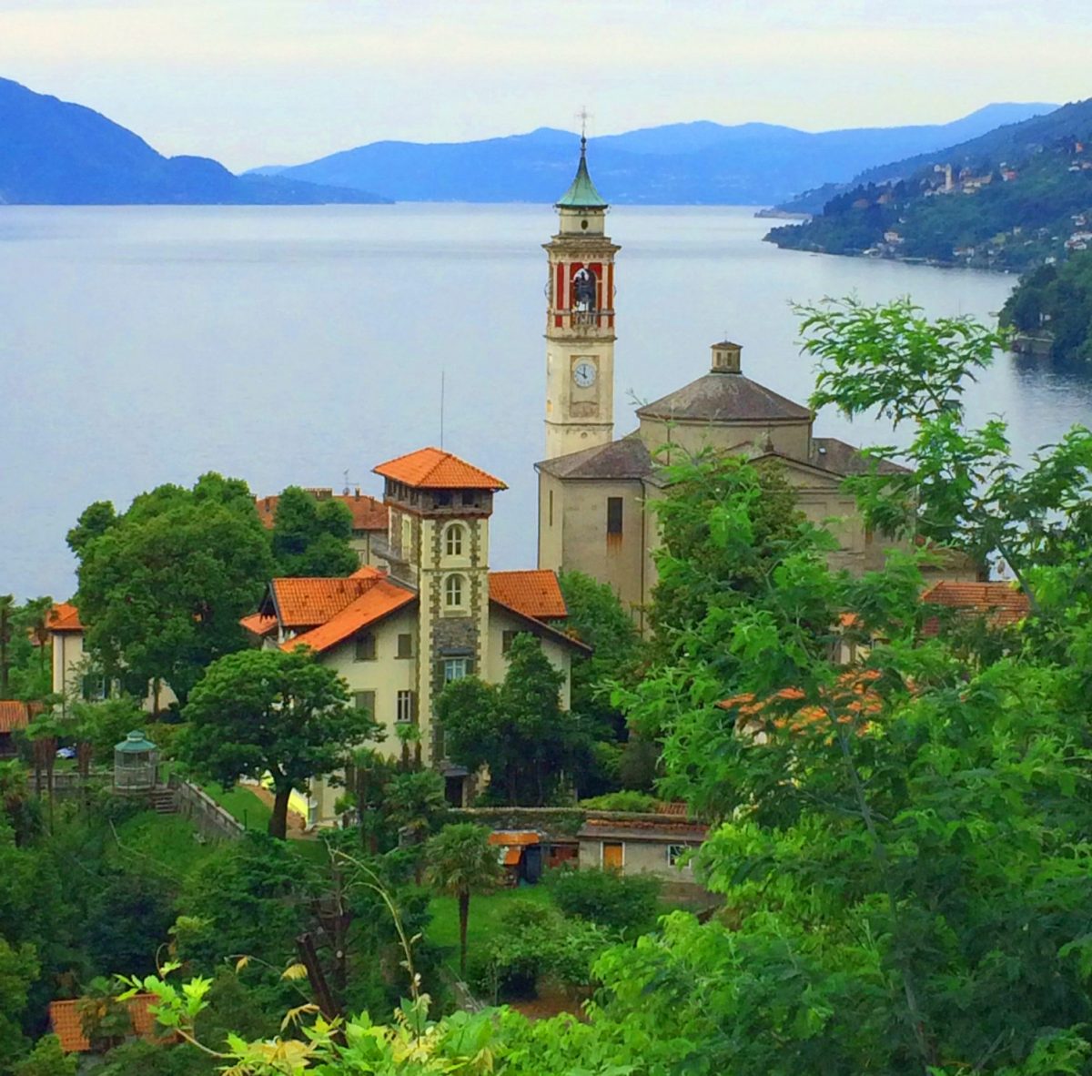 Scenic view of a lakeside village with a church tower, surrounded by lush greenery and mountains in the background.