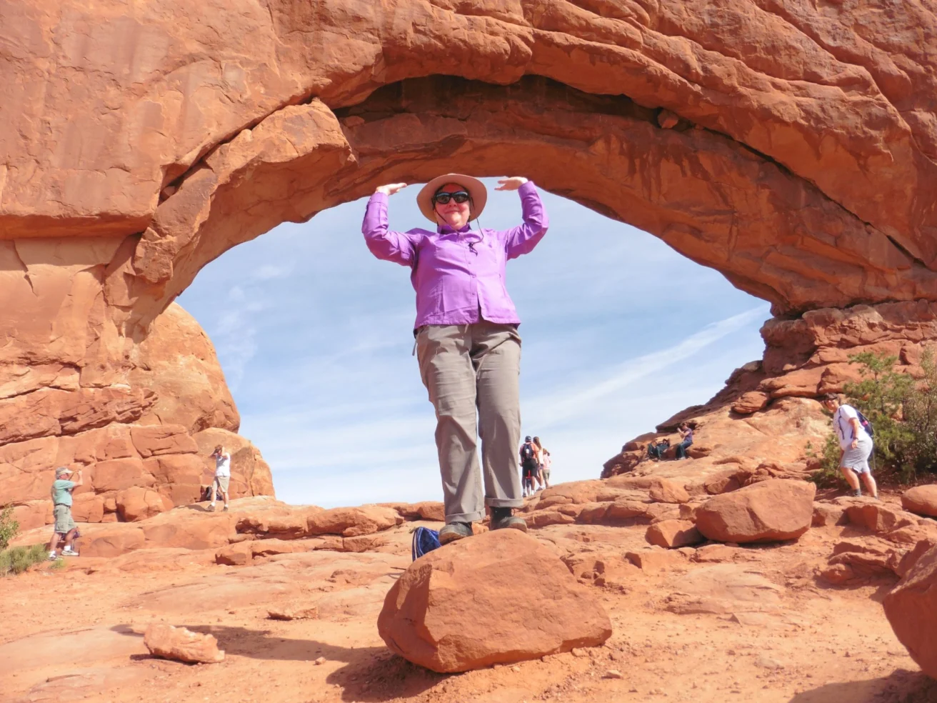 Person in a purple shirt and hat standing on a rock under a natural stone arch in a desert landscape. Other people are visible in the background.