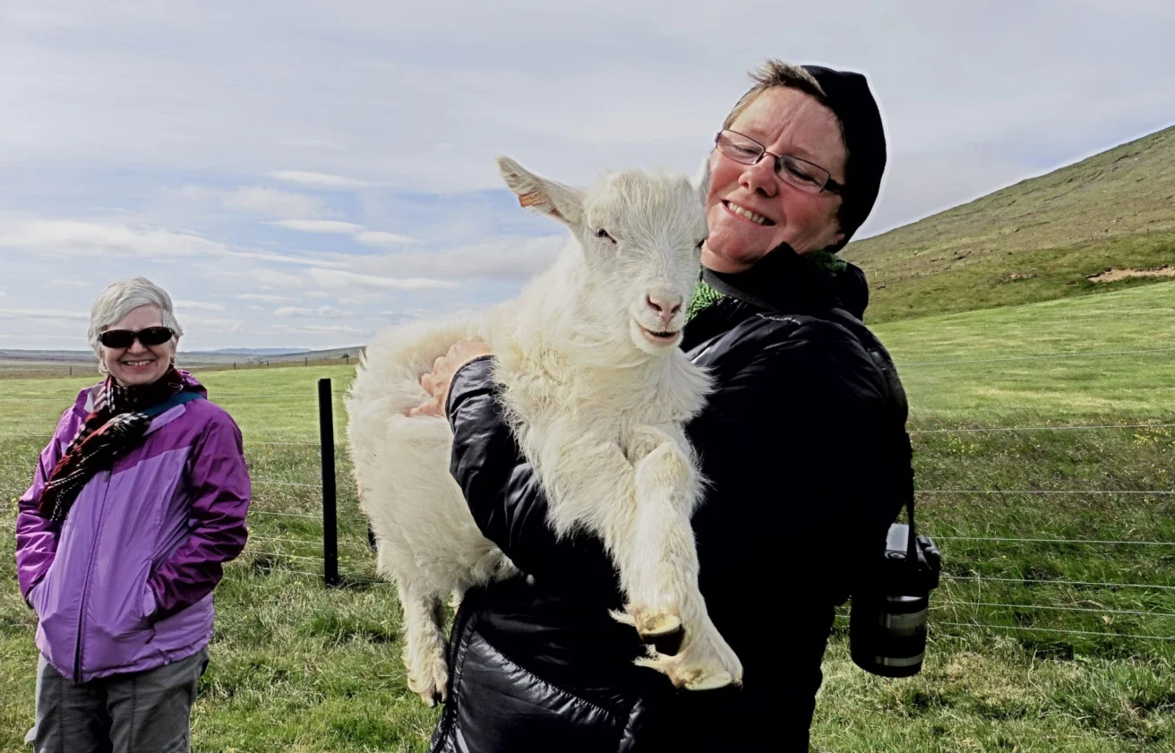 Two people in outdoor clothing, one holds a white goat and smiles. They're standing in a grassy area with a fence and hills in the background.