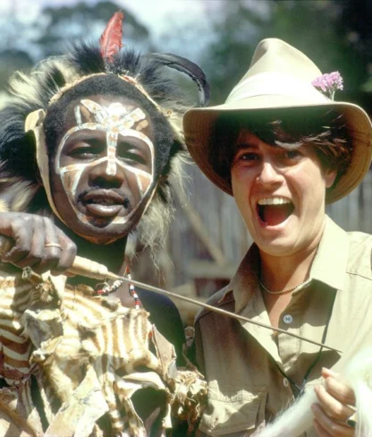 Person in traditional attire and face paint stands beside another person wearing a safari hat, both smiling at the camera, outdoors.