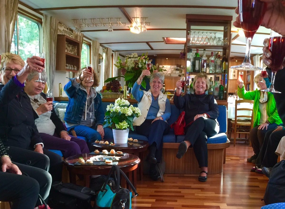 A group of people sitting in a room, raising glasses in a toast. There are flowers on a table and snacks are visible.