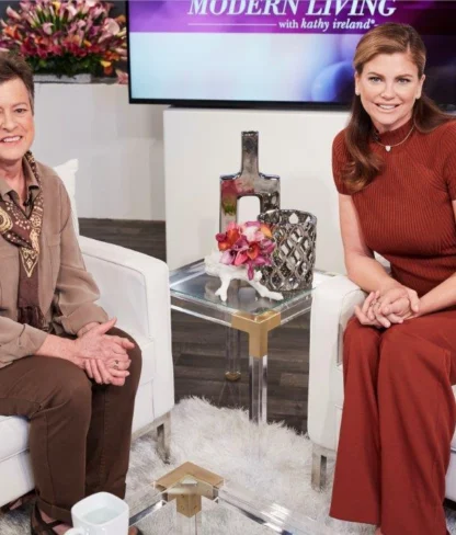 Two women sit in a TV studio. One is wearing a brown outfit with a scarf, and the other is in a rust-colored dress. They are seated on white chairs with decorative items around.