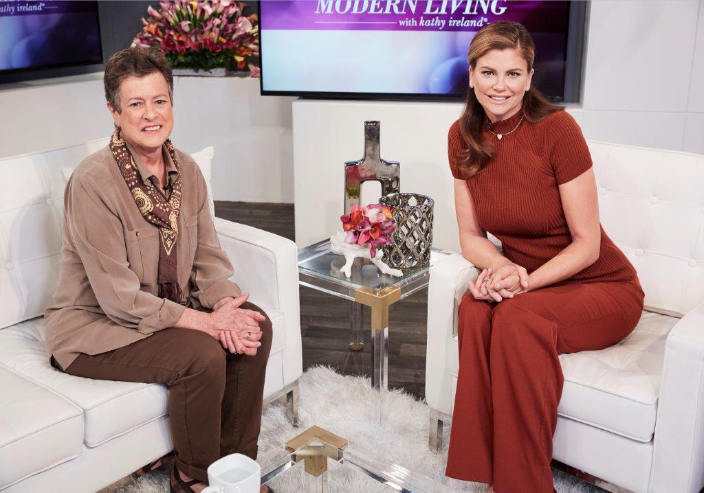 Two women sit in a TV studio. One is wearing a brown outfit with a scarf, and the other is in a rust-colored dress. They are seated on white chairs with decorative items around.