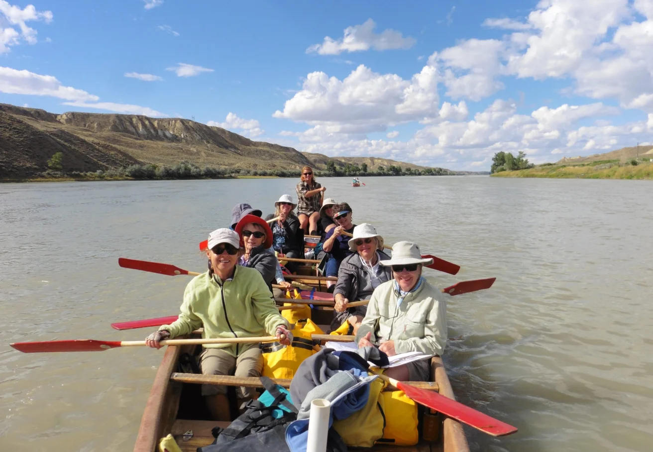 A group of people paddle a canoe on a wide river under a cloudy sky, surrounded by distant hills.