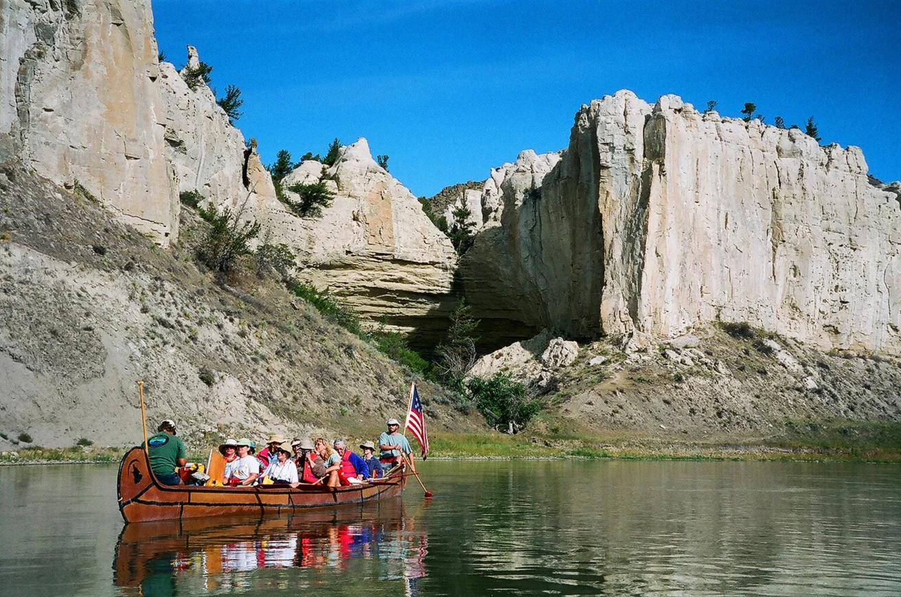 A group of people in a canoe with an American flag paddles on a river beside tall, rocky cliffs under a clear blue sky.