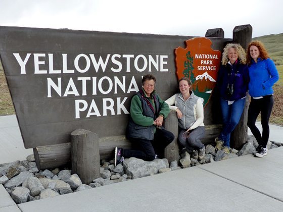 Four people pose by the Yellowstone National Park entrance sign, standing on a rocky surface.