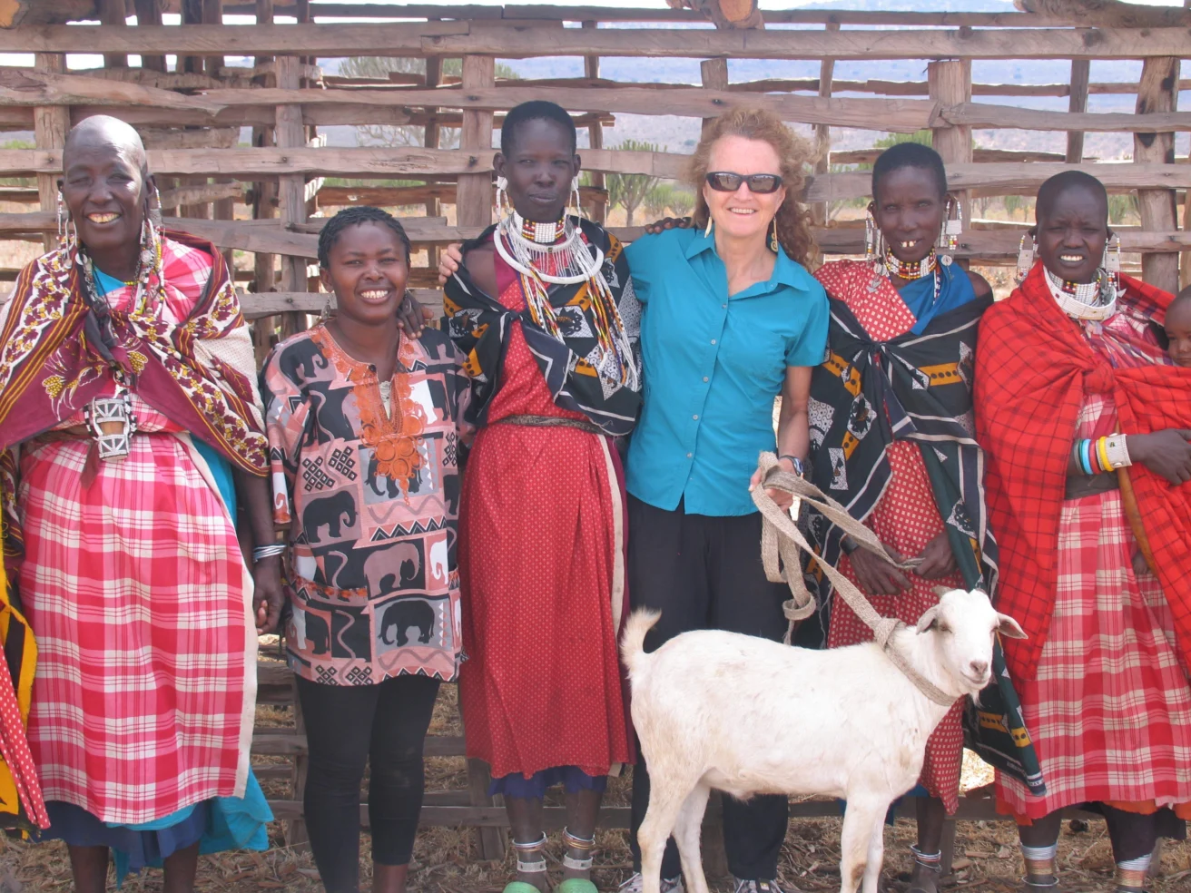 A group of women standing together with a goat in front of a wooden fence. They are wearing colorful clothing and accessories. One woman is wearing sunglasses.