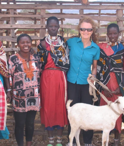 A group of women standing together with a goat in front of a wooden fence. They are wearing colorful clothing and accessories. One woman is wearing sunglasses.