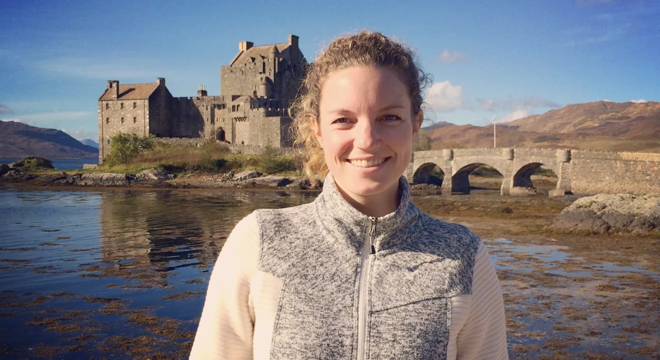 A person smiles in front of a historic stone castle situated by the water, with a bridge connecting to the castle.