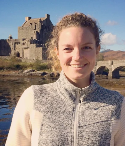 A person smiles in front of a historic stone castle situated by the water, with a bridge connecting to the castle.