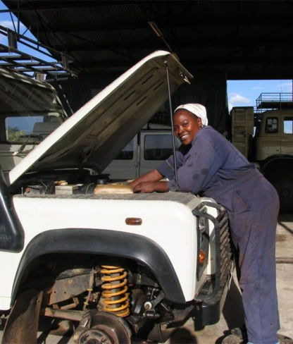 A mechanic wearing a headscarf works on the engine of a white vehicle inside a garage. Another vehicle is visible in the background.
