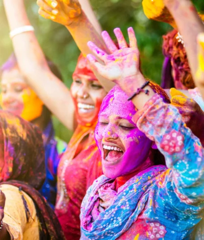 A group of people joyfully celebrating Holi, covered in colorful powders, with arms raised and smiling.