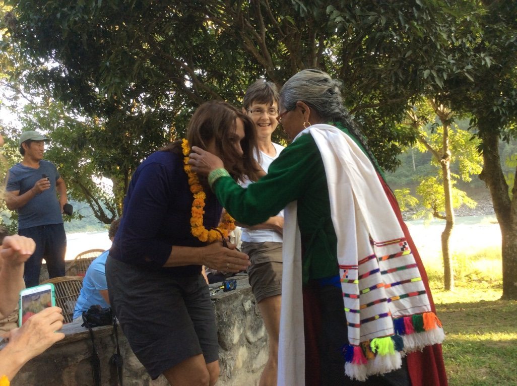 Person in traditional attire puts a flower garland on another person outdoors, with people watching in the background.