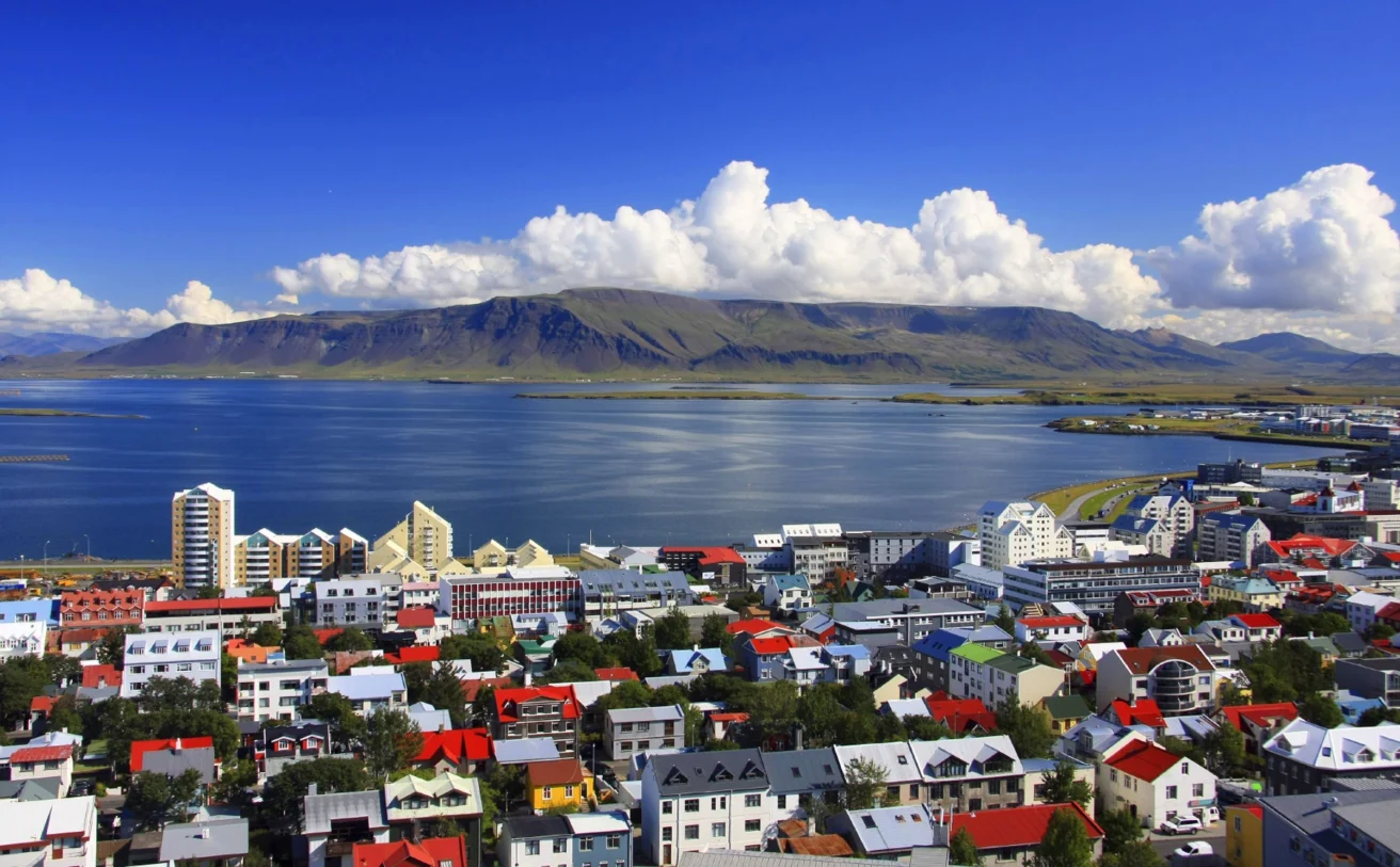 Colorful rooftops of a coastal town with a scenic view of a mountain range and a cloudy blue sky in the background.