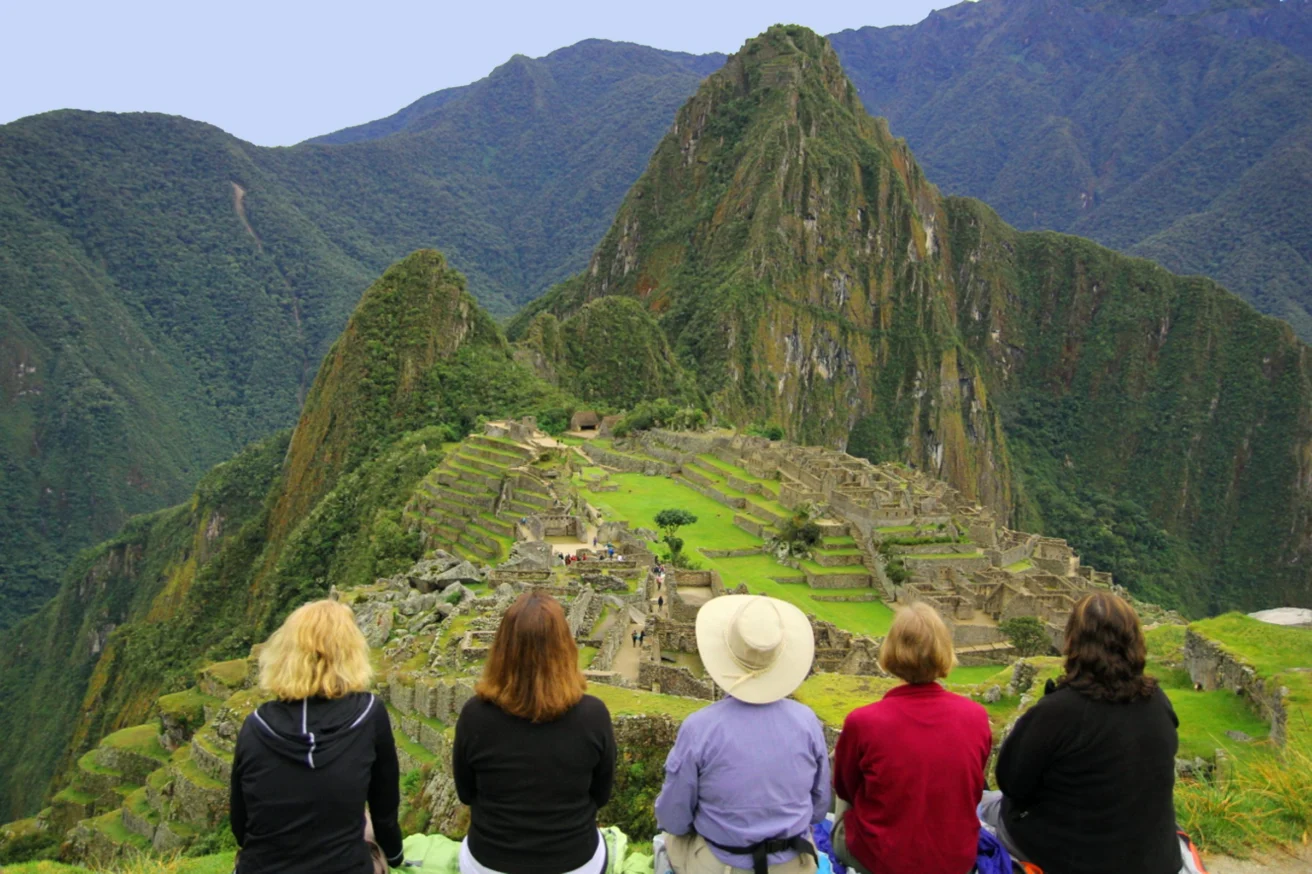 Five people sit and face the ancient Incan ruins of Machu Picchu, surrounded by lush green mountains under a clear sky.