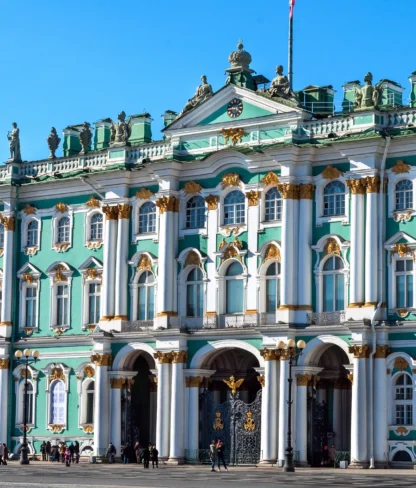 The image shows the Winter Palace in St. Petersburg, Russia, with its ornate green and white facade, columns, and statues under a clear blue sky.