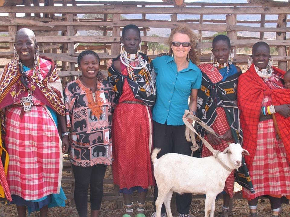 A group of people, including a woman in a blue shirt, stand together outdoors with a white goat. They are wearing colorful clothing and smiling. A wooden fence is in the background.