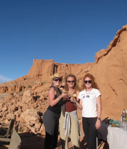 Three people standing outdoors near rock formations, holding drinks and smiling. A table with bottles is nearby under a clear blue sky.