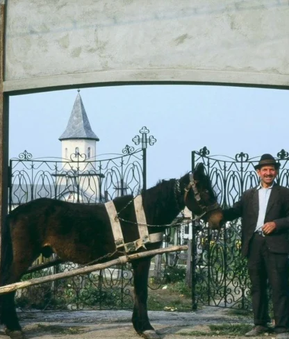 A man stands beside a horse-drawn cart under an archway, with a church steeple visible in the background.