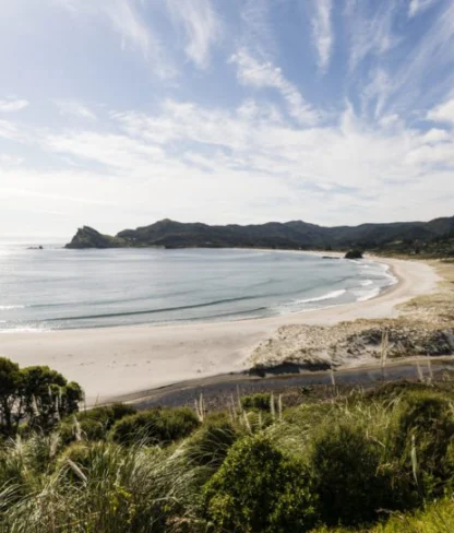 A scenic beach with a curving shoreline, sandy shores, and surrounding greenery under a partly cloudy sky.